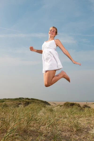 Pretty girl with red long hair wearing white dress enjoying nature near the beach. Jumping in the air. Hot summer day with blue cloudy sky. — Stock Photo, Image