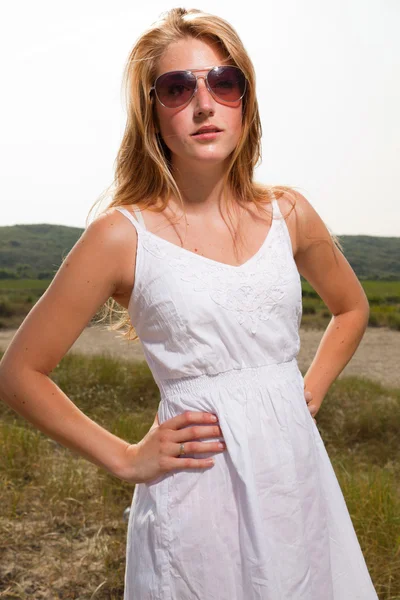 Красивая девушка с рыжими длинными волосами в белом платье и солнечных очках наслаждается природой рядом с пляжем. Жаркий летний день с голубым облачным небом . — стоковое фото