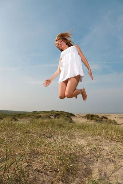 Chica bonita con el pelo largo rojo con vestido blanco disfrutando de la naturaleza cerca de la playa. Saltando en el aire. Día caluroso de verano con cielo azul nublado . —  Fotos de Stock