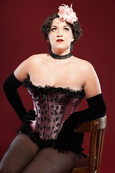 Sexy burlesque pin up mujer con el pelo largo rubio vestido de rosa y negro. Sentado en una silla de madera retro. Estudio de la moda tiro aislado sobre fondo rojo . —  Fotos de Stock