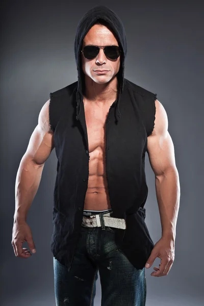 Muscled fitness man. Cool looking. Tough guy. Blue eyes. Blond short hair. Wearing black hoody shirt and sunglasses. Tanned skin. Studio shot isolated on grey background. — Stock Photo, Image