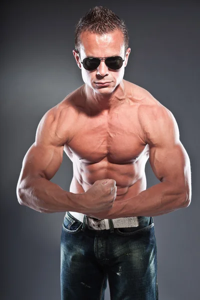 Shirtless muscled fitness man. Cool looking. Tough guy. Blue eyes. Blond short hair. Wearing black sunglasses. Tanned skin. Studio shot isolated on grey background. — Stock Photo, Image