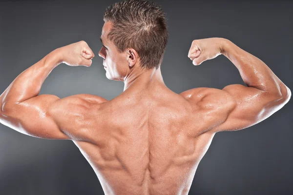 Shirtless muscled fitness man showing his back. Cool looking. Tough guy. Blue eyes. Blond short hair. Tanned skin. Studio shot isolated on grey background. — Stock Photo, Image