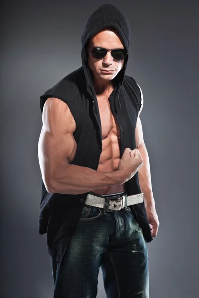 Muscled fitness man. Cool looking. Tough guy. Blue eyes. Blond short hair. Wearing black hoody shirt and sunglasses. Tanned skin. Studio shot isolated on grey background. — Stock Photo, Image