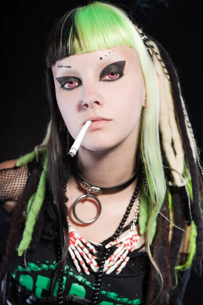 Cyber-Punk-Mädchen mit grün-blonden Haaren und roten Augen auf schwarzem Hintergrund. Gesichtsausdruck. Zigarette rauchen. Studioaufnahme. — Stockfoto