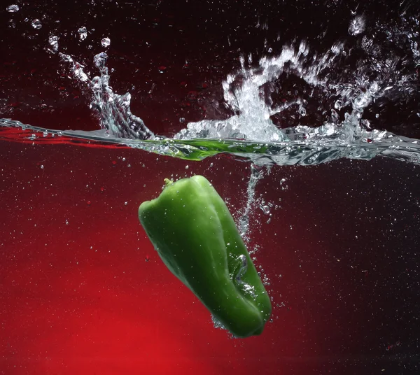 Grønn pepper som faller i vannet. Rød bakgrunn – stockfoto