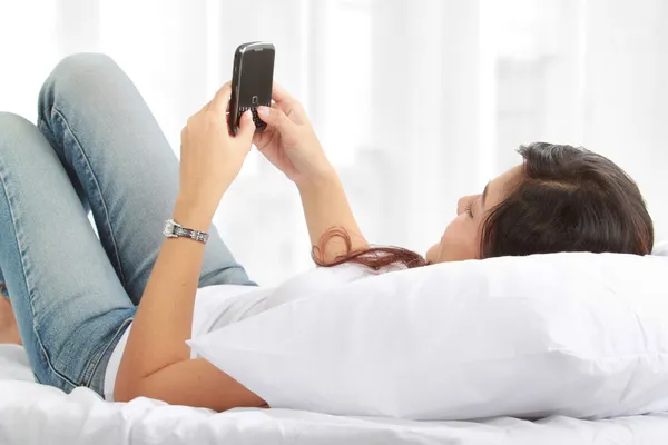 Žena SMS na telefonu ležící na posteli — Stock fotografie