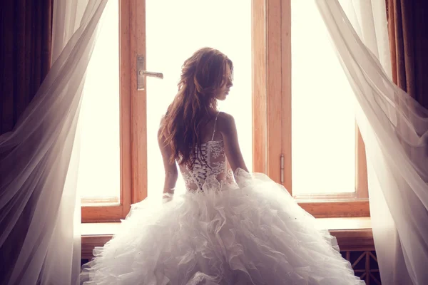 De mooie bruid tegen een raam binnenshuis Stockfoto