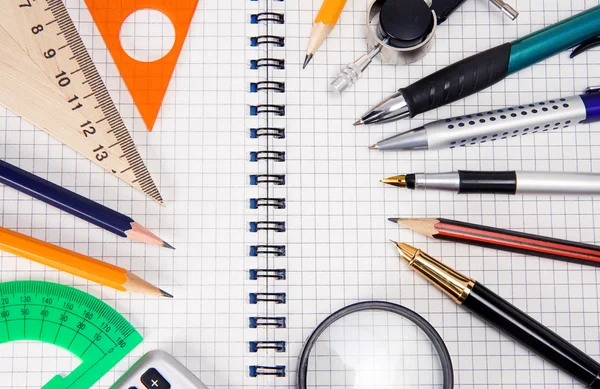 Pennen, potloden, Vergrootglas op cheked notebook — Stockfoto