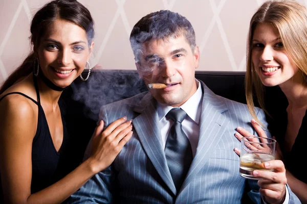 Retrato de un hombre exitoso fumando un cigarro sosteniendo whisky con mujeres bonitas cerca —  Fotos de Stock