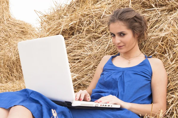 Brunette girl in blue dress typing on laptop on haystack — Stock Photo, Image