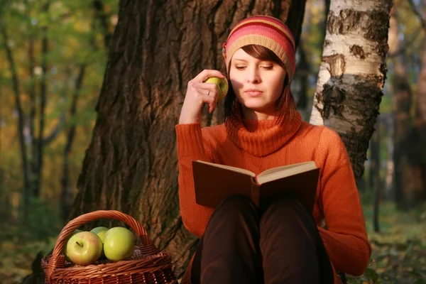 Young woman holding apple and reading book in fall park — Stock Photo, Image