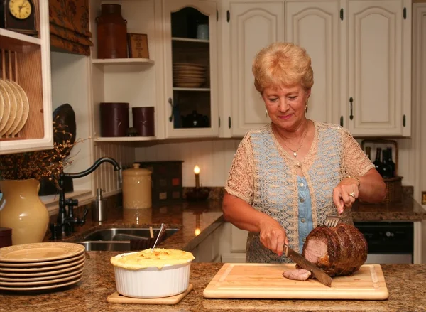 Grand-ma in a country kitchen carving a beef roast — Stock Photo, Image