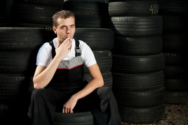 Jovem mecânico sentado no pneu e fumando — Fotografia de Stock
