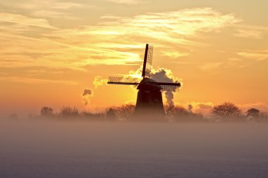 Traditional windmill in snow and fog in the countryside from the Netherlands at sunset clipart
