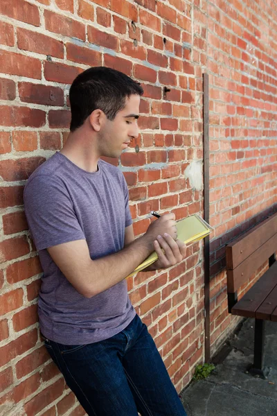 Bonito homem escrevendo em papel almofada por parede de tijolo — Fotografia de Stock