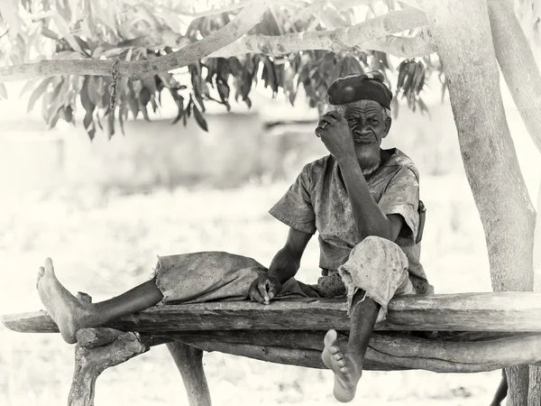 Um velho de Gana senta-se a maneira curiosa em um banco de madeira — Fotografia de Stock
