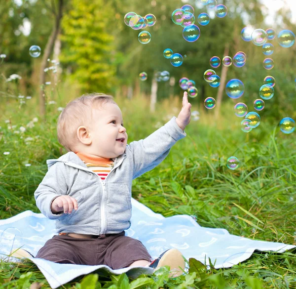 Menino sentado na grama verde outdor jogando com bolhas de sabão — Fotografia de Stock