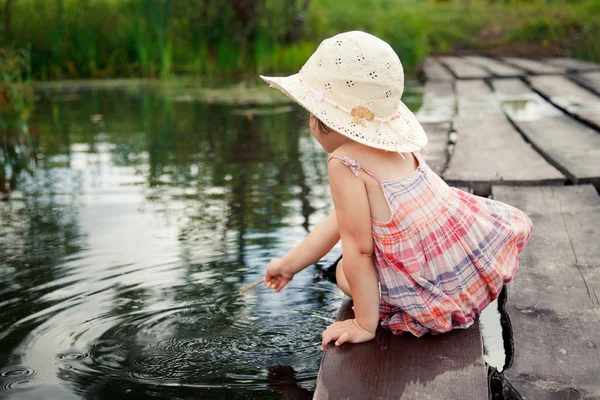 Entzückendes kleines Mädchen, das auf der Holzbrücke sitzt und nachdenklich auf den Fluss blickt — Stockfoto