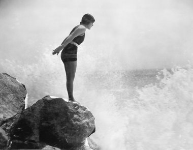 Female swimmer on rock above crashing surf clipart