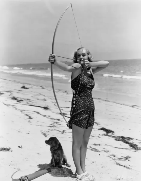 Woman shooting bow and arrow on beach — Stock Photo, Image
