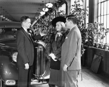 Man and woman standing in a car showroom talking to a salesman clipart