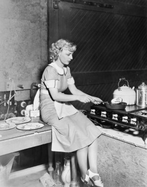 Young woman sitting on the counter and cooking food in the kitchen clipart