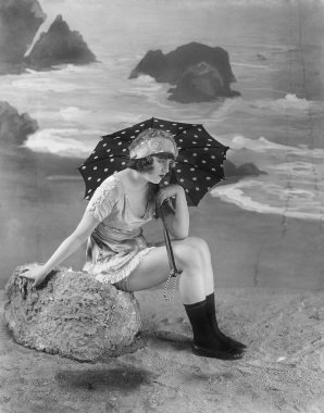 Young woman sitting on a bolder on the beach, holding an umbrella clipart
