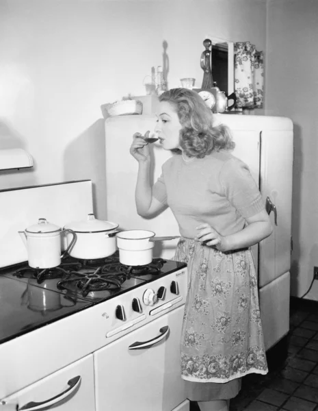 Mladá žena v zástěře v její kuchyni ochutnávka jí jídlo z hrnce — Stock fotografie