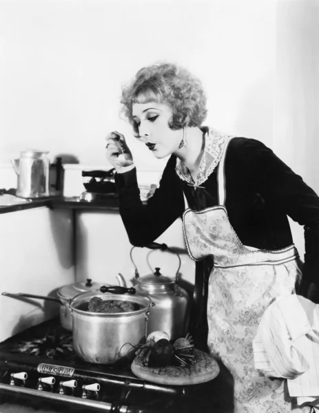 Νεαρή γυναίκα σε μια ποδιά στην κουζίνα της γεύση το φαγητό από ένα δοχείο — Φωτογραφία Αρχείου