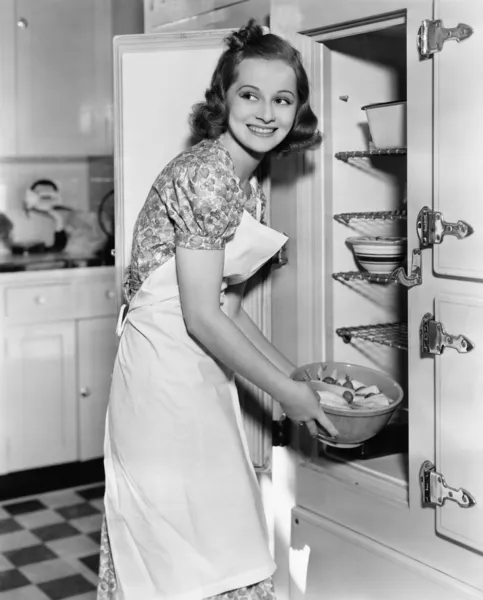 Young woman in an apron in her kitchen taking food out of the refrigerator — Stock Photo, Image