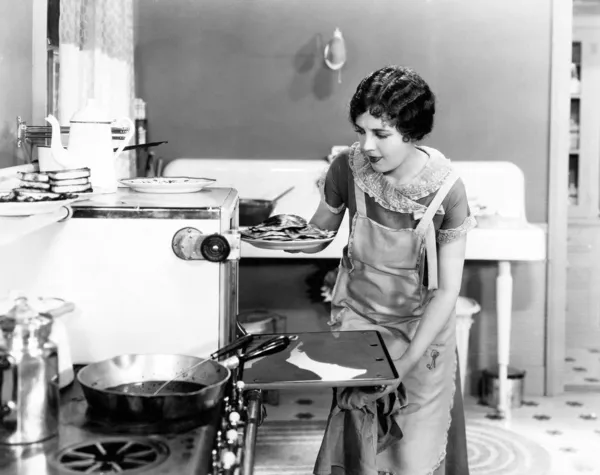 Jovem mulher em um avental em sua cozinha tirando comida do forno — Fotografia de Stock