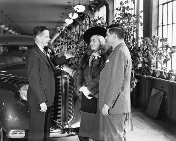Man and woman standing in a car showroom talking to a salesman — Stock Photo, Image