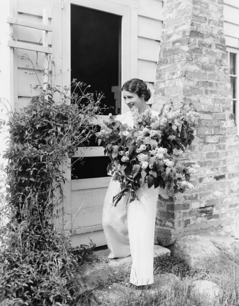Γυναίκα μπροστά από το σπίτι που συλλέγουν τα λουλούδια στην αγκαλιά της — Φωτογραφία Αρχείου