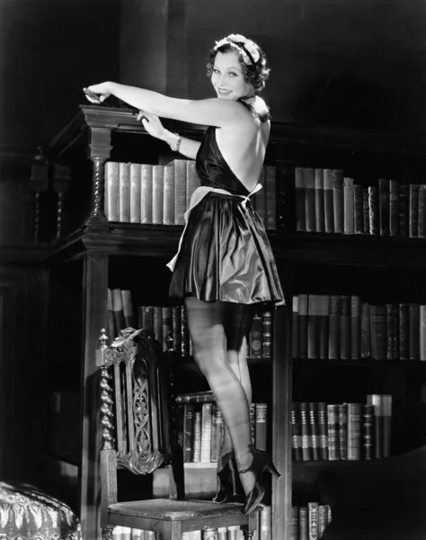 Retrato de una mujer joven de pie en una silla y desempolvar una estantería en un traje sexy —  Fotos de Stock