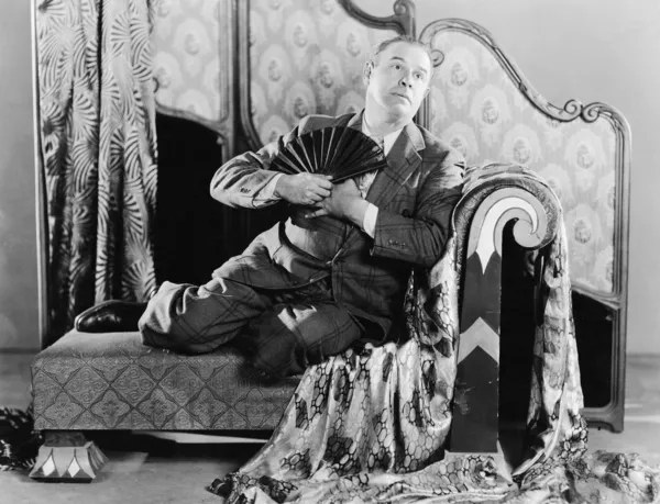 Retrato de un hombre sentado en un sofá sosteniendo un ventilador — Foto de Stock
