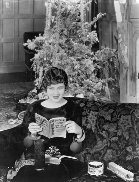 Mujer joven sentada en un sofá con un árbol de Navidad en el fondo — Foto de Stock