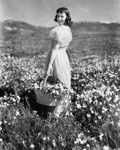 Вид сзади на девушку, стоящую на лугу с корзиной цветов и улыбающуюся — стоковое фото