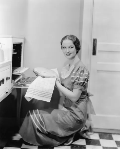 Профиль молодой женщины, достающей пирог из духовки, улыбающейся в камеру — стоковое фото