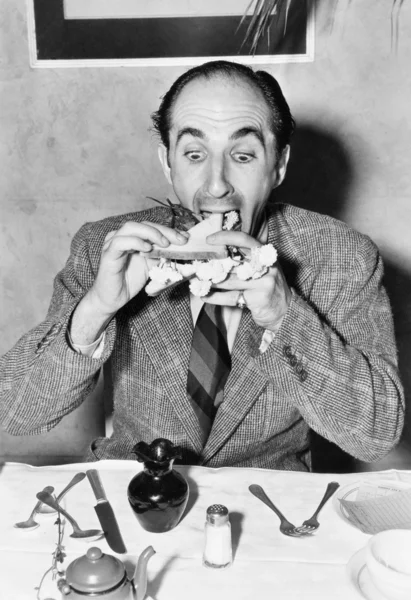 Человек, сидящий за обеденным столом и пытающийся съесть сэндвич — стоковое фото