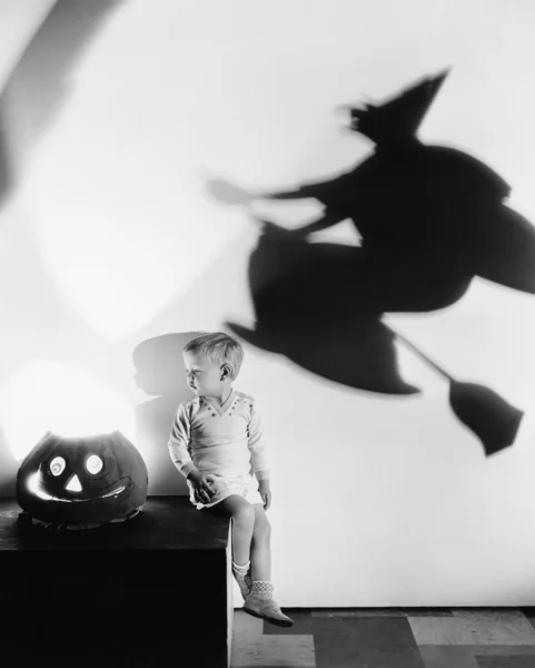 Çocuk bir masada oturuyor, balkabağı ve duvarda bir cadının gölgesi var. — Stok fotoğraf