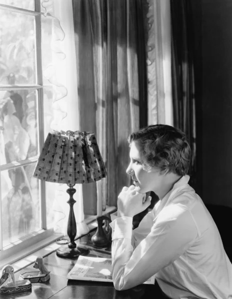 Profil av en ung kvinna sitter vid ett skrivbord och titta igenom ett fönster — Stockfoto