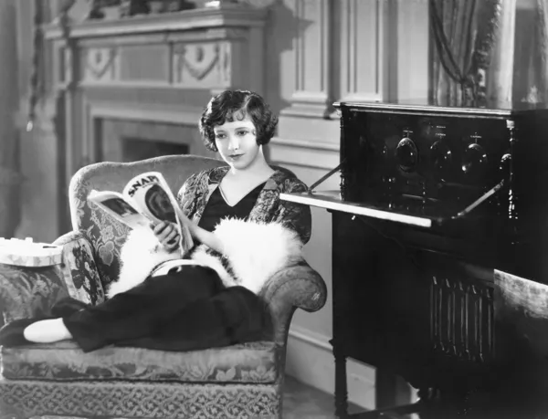 Ung kvinna sitter i en fåtölj som läser en tidning vid en radio — Stockfoto