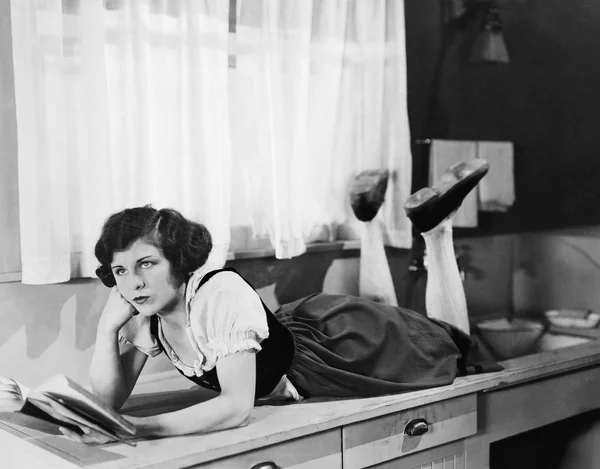 Mujer joven acostada en un mostrador de cocina sosteniendo un libro y pensando —  Fotos de Stock