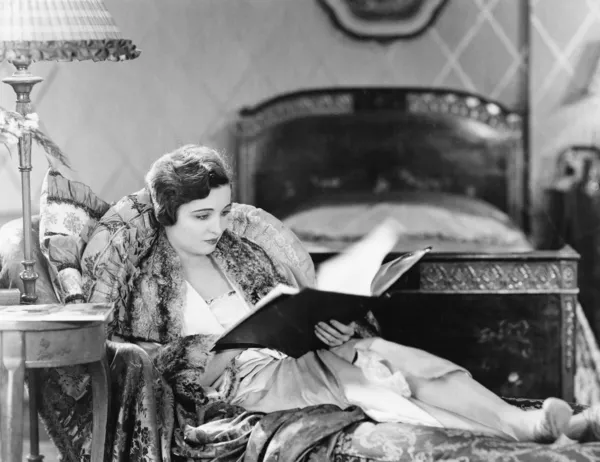 Junge Frau legt sich in einen Sessel und liest ein Buch in ihrem Schlafzimmer — Stockfoto