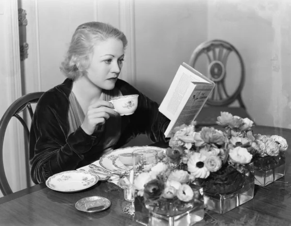 Jovem sentada na mesa de jantar segurando uma xícara de chá e lendo um livro — Fotografia de Stock