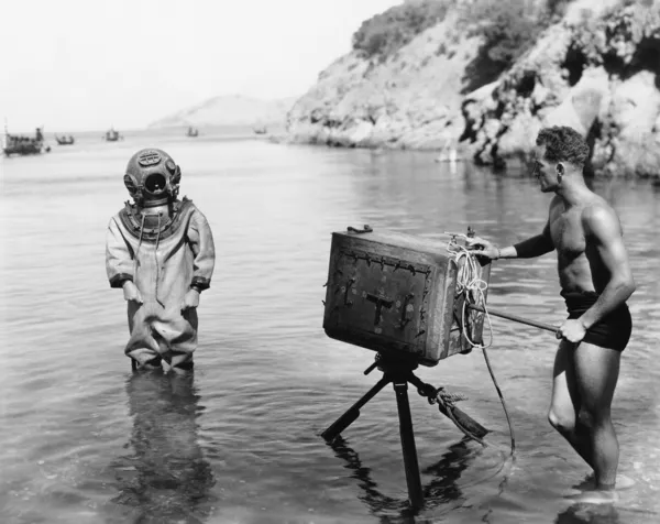 Профиль молодого человека, держащего камеру с аквалангистом, стоящим перед ним на пляже — стоковое фото