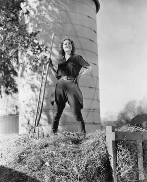 Νεαρή γυναίκα στέκεται πάνω σε μια θυμωνιά χόρτου και κρατώντας ένα pitchfork — Φωτογραφία Αρχείου