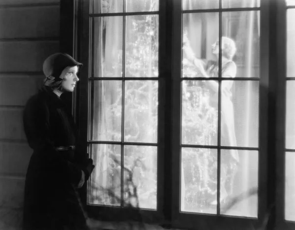 Bir kadın bir Noel ağacı süsleme izlerken bir pencere dışında duran kadın — Stok fotoğraf