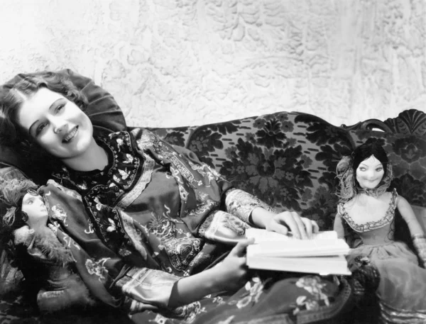Mulher em um vestido de impressão chinesa deitada no sofá com suas bonecas e um livro — Fotografia de Stock